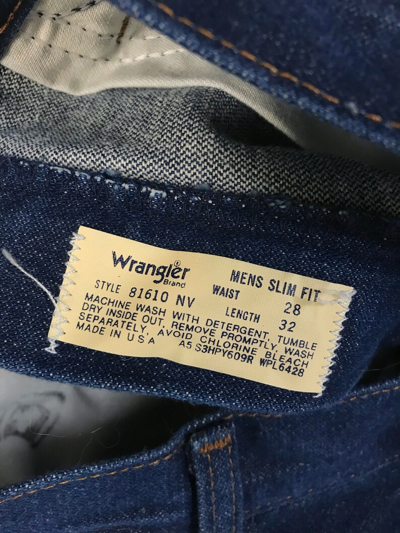 May include: A close-up of a blue denim Wrangler jeans label with white text. The label states "Style 81610 NV", "Mens Slim Fit", "Waist", "Length", "28", "32", "Machine Wash with Detergent, Tumble Dry Inside Out, Remove Promptly, Wash Separately, Avoid Chlorine Bleach", "Made in USA", "A5 S3HPY609R WPL6428".