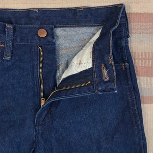 May include: A pair of dark blue denim jeans with a zipper fly and a brown button closure. The jeans are open to show the inside of the waistband.