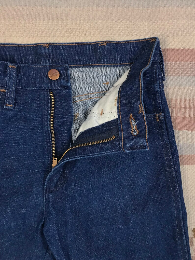May include: A pair of dark blue denim jeans with a zipper fly and a brown button closure. The jeans are open to show the inside of the waistband.