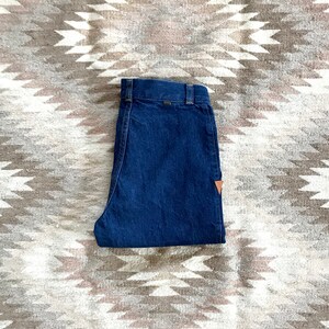 Op de afbeelding: Opgevouwen donkerblauwe jeansbroek met een bruin leren patch op de achterzak. De broek ligt op een kleed met een geometrisch patroon.