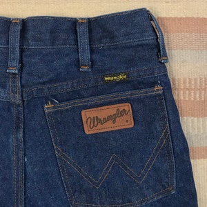 May include: Dark blue denim jeans with a brown leather Wrangler patch on the back pocket.