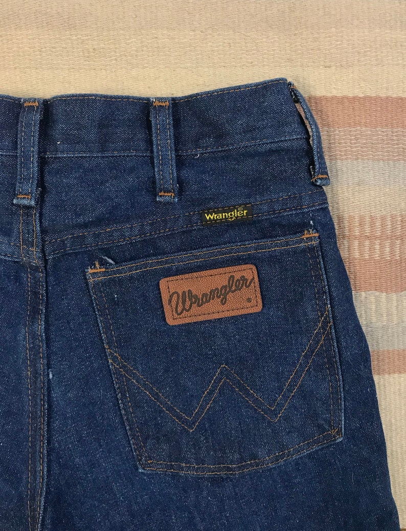 May include: Dark blue denim jeans with a brown leather Wrangler patch on the back pocket.