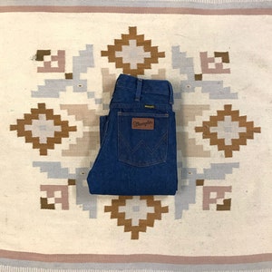 May include: A pair of blue denim jeans folded on a white and brown patterned rug. The jeans have a brown leather patch with the word "Wrangler" on it.