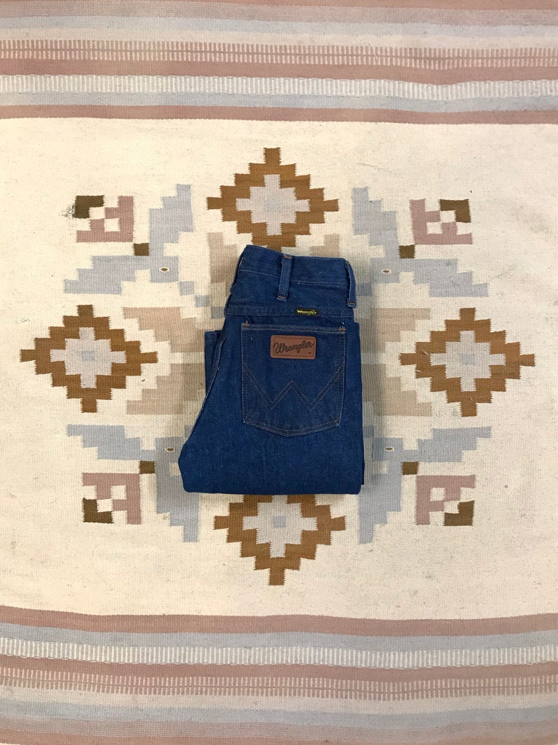 May include: A pair of blue denim jeans folded on a white and brown patterned rug. The jeans have a brown leather patch with the word "Wrangler" on it.