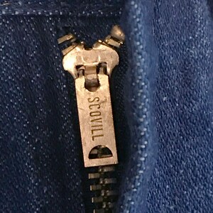 May include: Close-up of a silver zipper pull with the word "SCOVILL" engraved on it. The zipper is on a pair of blue denim jeans.