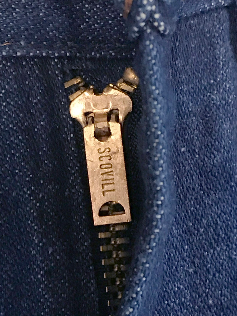 May include: Close-up of a silver zipper pull with the word "SCOVILL" engraved on it. The zipper is on a pair of blue denim jeans.