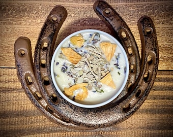 May include: A white candle with dried herbs and flowers sits inside a bronze horseshoe. The candle is surrounded by a bronze horseshoe on a wooden surface. The candle has a creamy white wax with dried herbs and flowers.