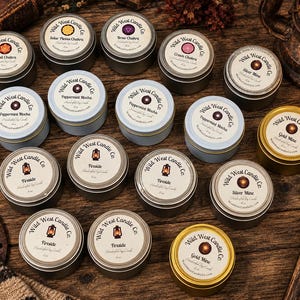 May include: A collection of round metal candle tins with labels reading Wild West Candle Co. The labels feature various scents and themes including Sacral Chakra, Solar Plexus Chakra, Brow Chakra, Crown Chakra, Silver Mine, Peppermint Mocha, Fireside, and Gold Mine.