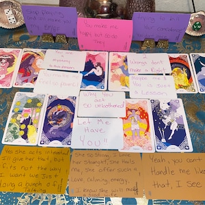 May include: A tarot card reading spread with handwritten notes on cards and paper. The cards are on a blue patterned surface. The notes include phrases like "You make me happy but so do they", "Why you act so unbothered", and "Let me have you!"