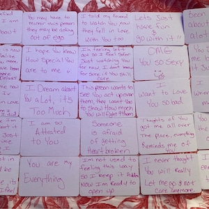 May include: A set of 12 pink cards with handwritten messages about love and relationships. The cards are arranged in a grid on a red and white patterned surface.