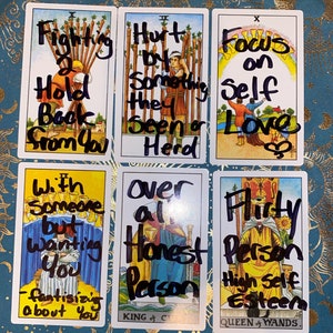 May include: Six tarot cards with handwritten text. The cards feature illustrations and phrases like "Fighting Hold Back From You," "Hurt by Something They Seen a Herd," and "Focus on Self Love." The cards are arranged on a teal surface.