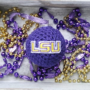 May include: A purple crocheted ornament with the letters "LSU" in yellow, surrounded by gold and purple beaded necklaces. The ornament is in a white wooden tray with rope handles.