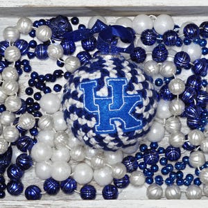 May include: A decorative ornament with a blue and white knitted pattern, featuring a blue "UK" logo. The ornament is surrounded by strands of white and blue beads, and a blue ribbon. The arrangement is set in a white tray.