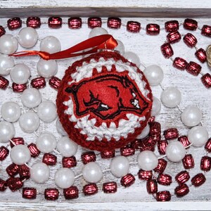 May include: A red and white crocheted Christmas ornament featuring a Razorback logo, with a red ribbon for hanging. The ornament is surrounded by strings of white pearl beads and red, rectangular beads, all arranged in a white wooden tray.