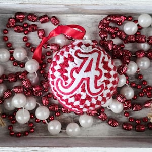 May include: A decorative Christmas ornament with a red and white pattern and a large white letter "A" is displayed in a wooden tray. The ornament is surrounded by red beaded garland and white pearl beads, creating a festive holiday scene.