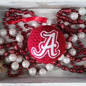May include: A red crocheted Christmas ornament with a white "A" logo, surrounded by red and white beaded garlands, and a red ribbon. The ornament and garlands are arranged in a white wooden tray.