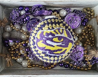 May include: A decorative Christmas ornament with a purple and yellow design, surrounded by purple, gold, and white beads and purple coins. The ornament features a logo. The items are arranged in a wooden tray.