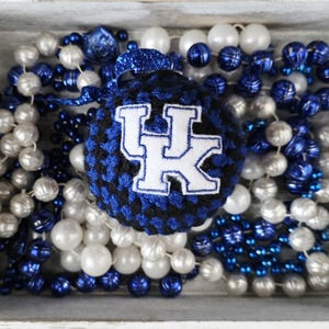 May include: A blue and black crocheted ornament with the letters "UK" in white, surrounded by blue, silver, and white beaded necklaces. The ornament is in a wooden tray with rope accents.