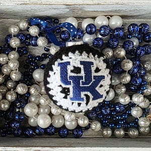 May include: A handmade Christmas ornament with a black crocheted base and a white and blue UK logo. The ornament is surrounded by blue, silver, and white beaded garlands, and a blue ribbon. The ornament is in a wooden tray.