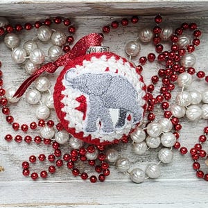 May include: A red and white crocheted Christmas ornament with a gray elephant design. The ornament is surrounded by red and white beaded garlands in a white wooden tray. Decorative holiday item.