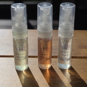 May include: Three clear perfume sample bottles with spray tops. The bottles contain different colored liquids and are labeled "Laundry Room", "Essence of Kesh", and "Peachy Peach". The bottles are on a wooden surface.