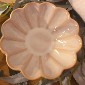 May include: A small, light pink ceramic dish with a scalloped edge. The dish has a flower-like shape with a lighter center and darker pink accents. The dish is likely used for serving small portions of food or as a decorative item.