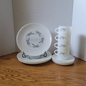 May include: A set of white vintage dinnerware with a wheat design. The set includes a large plate, a smaller plate, and four cups.