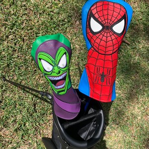 May include: Two golf club head covers featuring comic book characters. One cover is green and purple, depicting a grinning villain. The other is red and blue, with a spider web design and a black spider emblem.