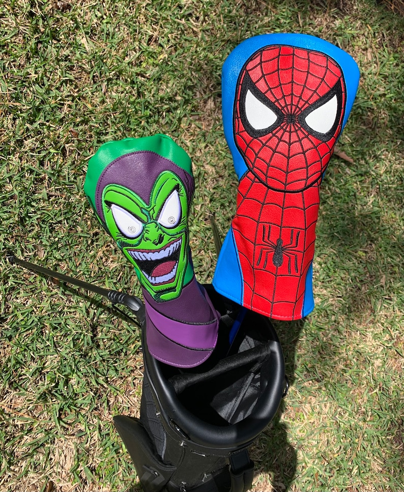 May include: Two golf club head covers featuring comic book characters. One cover is green and purple, depicting a grinning villain. The other is red and blue, with a spider web design and a black spider emblem.