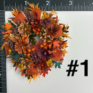 May include: A circular autumn-themed wreath decoration featuring orange burlap ribbon, brown pinecones, dark red berries, and assorted fall-colored leaves. A black ruler is visible along the top and left edges, showing the item is 3 inches wide.
