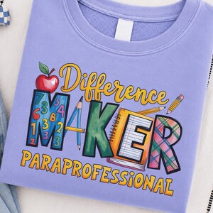 May include: A light purple crewneck sweatshirt featuring a graphic print with the text Difference Maker Paraprofessional. The design includes a red apple, a blue letter M filled with colourful numbers, a green letter A, a yellow notebook, and a plaid letter R.