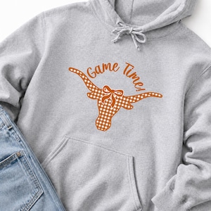 May include: A light gray hooded sweatshirt featuring a graphic of a longhorn steer head with a white and orange gingham pattern and a small bow between the horns. The text Game Time! is printed in orange above the longhorn graphic.