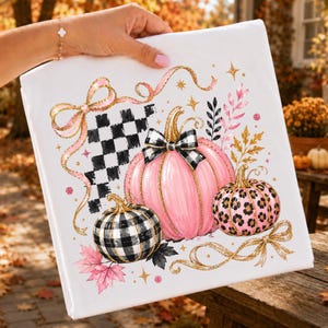 May include: A white fabric item featuring a graphic print of three pumpkins. One pumpkin is solid pink with a black and white checkered bow, one is black and white checkered, and one is pink with black leopard spots. Gold glitter ribbons and leaves.