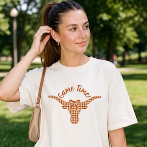 May include: A cream-colored short-sleeve t-shirt featuring a burnt orange graphic of a longhorn steer head with a bow between the horns. The steer head has a gingham check pattern, and the text "Game Time!" is printed above in orange.
