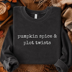 May include: A charcoal gray crewneck sweatshirt featuring the white text "pumpkin spice & plot twists" printed on the front. The garment has long sleeves, a ribbed collar, and a standard fit, showcasing a clean and simple design for casual wear.