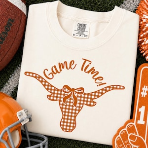 May include: A cream-colored t-shirt featuring the text Game Time! in orange script above a graphic of a longhorn steer head filled with an orange and white gingham pattern and accented with a small bow between the horns on the forehead.