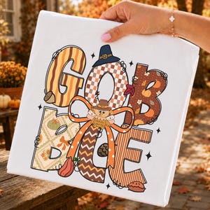 May include: A white t-shirt featuring the word GOBBLE in large, stylized letters. Each letter has a unique pattern including stripes, checkers, and autumn motifs. A cartoon turkey wearing a pilgrim hat sits in the center with an orange bow.