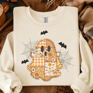 May include: A cream-colored crewneck sweatshirt featuring a central ghost graphic composed of patchwork patterns including orange plaid, autumn leaves, and floral prints. The design is accented by black spiderwebs and three small black bats.