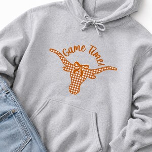 May include: A light gray hooded sweatshirt featuring a graphic of a longhorn steer head with a white and orange gingham pattern and a small bow between the horns. The text Game Time! is printed in orange above the longhorn graphic.