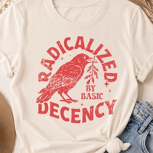 Op de afbeelding: Een crèmekleurig T-shirt met korte mouwen en een verweerde rode afbeelding van een kraai die een takje in de snavel houdt. De tekst RADICALIZED BY BASIC DECENCY is in een cirkelvormig, verweerd rood lettertype rond de vogel gedrukt.