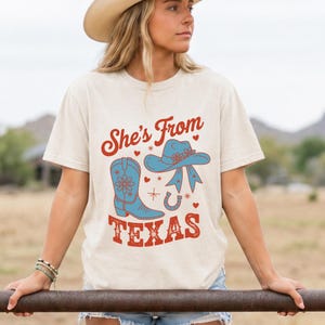 May include: A cream-colored short-sleeve t-shirt featuring a graphic print with the text "She's From TEXAS" in red. The design includes a blue cowboy boot, a blue cowboy hat with a ribbon, a horseshoe, small red hearts, and star accents.