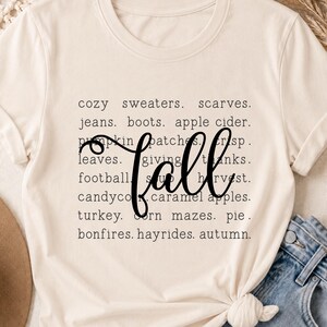 May include: A cream-colored short-sleeve t-shirt featuring black text listing fall-themed words like cozy sweaters, scarves, jeans, boots, apple cider, pumpkin patches, crisp leaves, giving thanks, football, soup, harvest, candy corn, caramel apples, turkey, corn mazes, pie, bonfires, hayrides, and autumn, with the word fall written in a large black script font across the center.