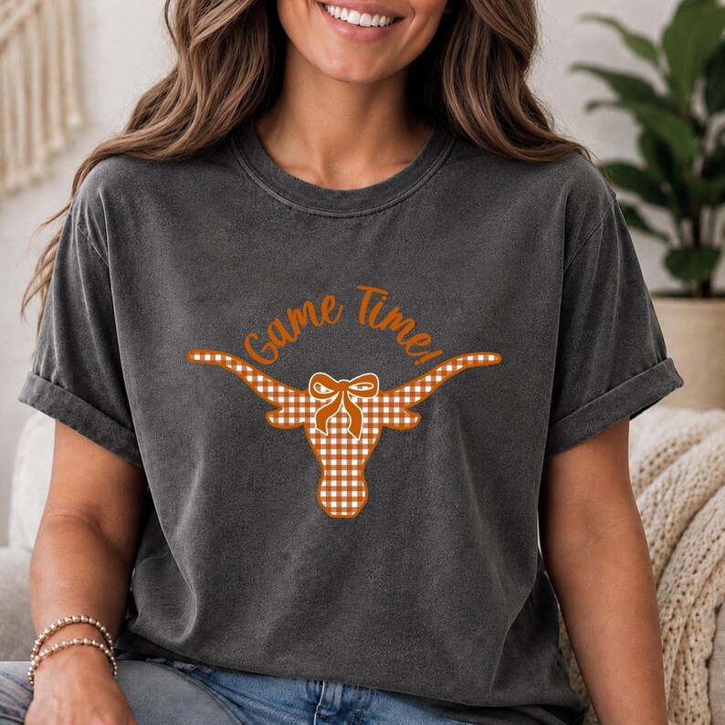 May include: A charcoal gray short-sleeve t-shirt featuring a graphic of a longhorn steer head with a bow, patterned in an orange and white gingham check. Above the steer head, the text Game Time! is printed in an orange script font.