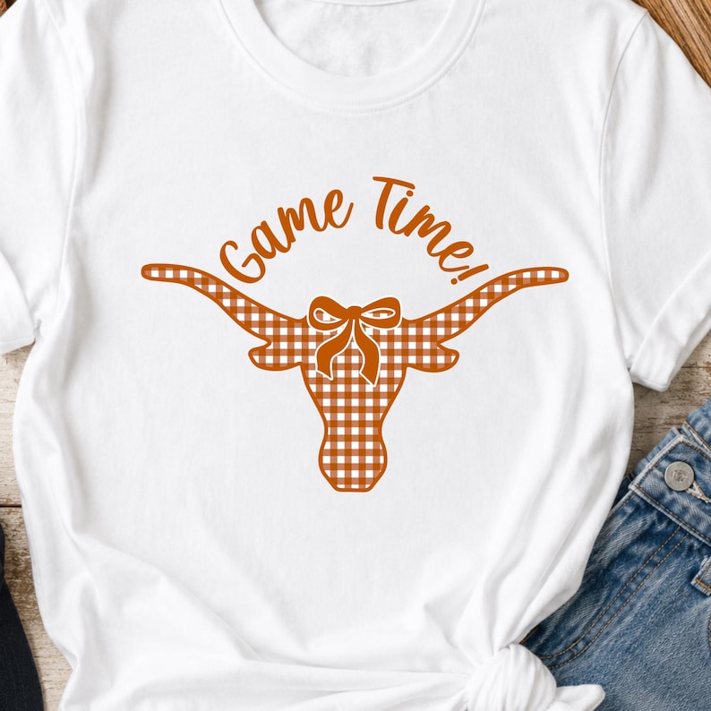 May include: A white short-sleeved t-shirt featuring a graphic of a longhorn steer head with a brown and white gingham pattern. The steer has a small bow between its horns, and the text Game Time! is printed in brown above the steer head.