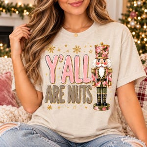 Puede incluir: Una camiseta beige de manga corta con un gráfico de un cascanueces con uniforme de camuflaje rosa y verde. El texto Y'ALL ARE NUTS está impreso en letras grandes rosas y grises, rodeado de pequeños copos de nieve y estrellas doradas brillantes.