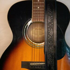 Puede incluir: Una guitarra ac&uacute;stica marr&oacute;n y negra con una correa de cuero negra. La guitarra tiene una boca de sonido con una etiqueta que dice "Mitchell, Modelo No. 100-GT, 6 Cuerdas".