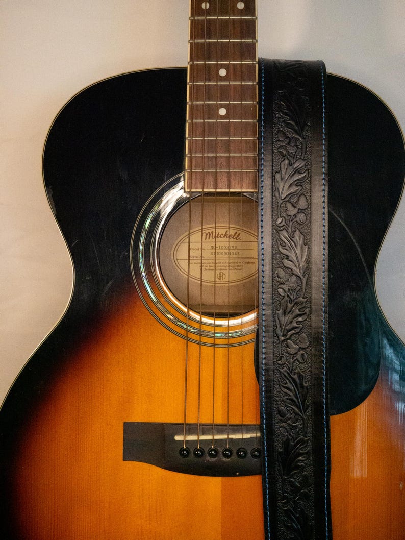 Puede incluir: Una guitarra ac&uacute;stica marr&oacute;n y negra con una correa de cuero negra. La guitarra tiene una boca de sonido con una etiqueta que dice "Mitchell, Modelo No. 100-GT, 6 Cuerdas".