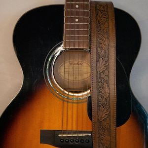 Puede incluir: Una guitarra ac&uacute;stica marr&oacute;n con una golpeador negro y una correa de guitarra de cuero marr&oacute;n con un dise&ntilde;o floral en relieve. La guitarra tiene una boca de sonido con una etiqueta que dice "Mitchell  MC-300CE  Made in China".