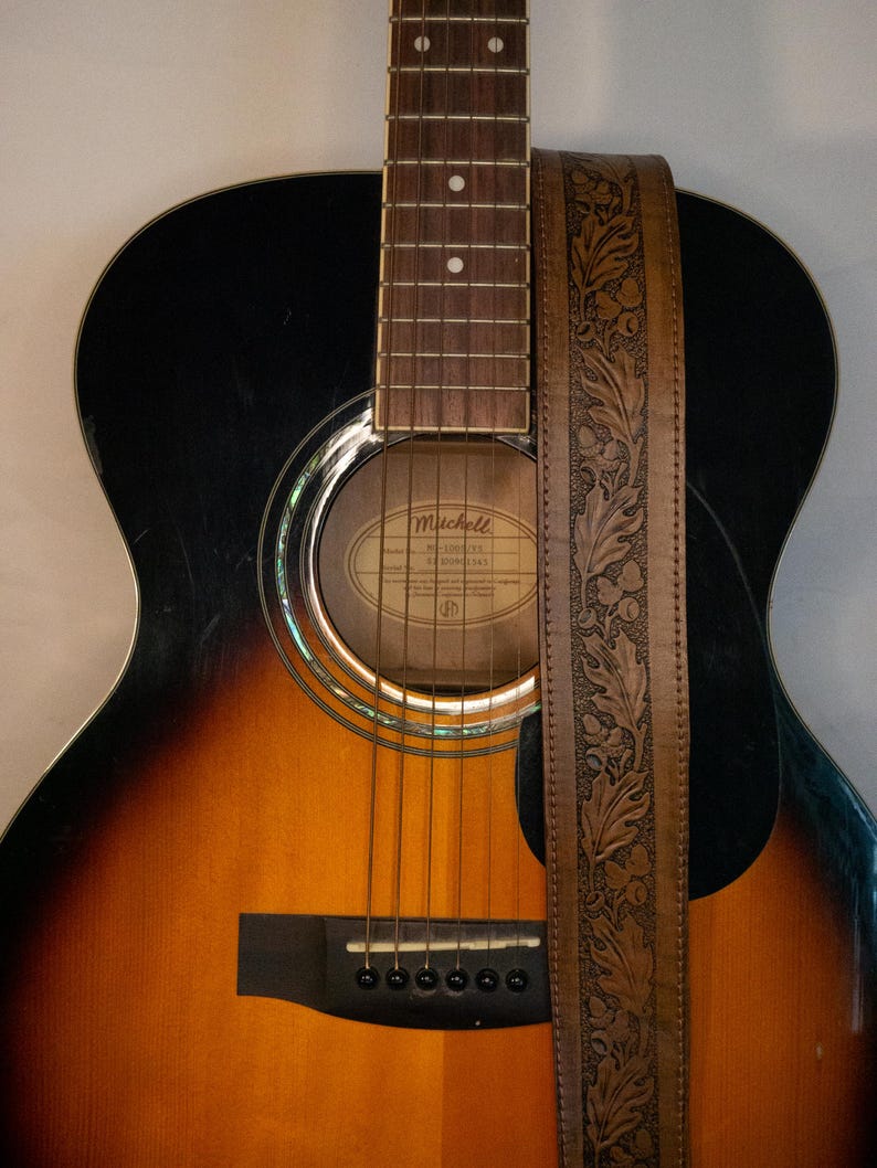 Puede incluir: Una guitarra ac&uacute;stica marr&oacute;n con una golpeador negro y una correa de guitarra de cuero marr&oacute;n con un dise&ntilde;o floral en relieve. La guitarra tiene una boca de sonido con una etiqueta que dice "Mitchell  MC-300CE  Made in China".