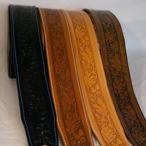 May include: Four leather guitar straps with an embossed floral design. The straps are in different shades of brown, with one being black. The straps have a white stitch detail.